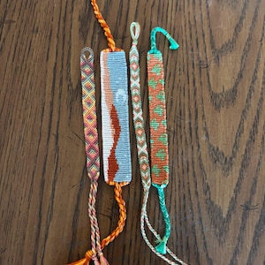 May include: Five colorful friendship bracelets with intricate woven patterns. The bracelets feature a variety of colors, including orange, red, blue, and green. Each bracelet has a unique design and is made with braided threads. The bracelets are displayed on a wooden surface.