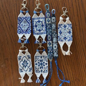 May include: A collection of six handmade woven items in shades of blue and white. Four items are rectangular keychains with silver metal lobster clasps, and two are long, narrow woven bracelets with braided ends featuring geometric patterns.