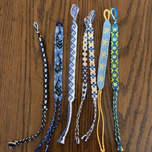 May include: Seven handmade friendship bracelets in various colours and patterns. The bracelets feature designs in blue, white, yellow, and orange, with braided and woven details. Each bracelet has a loop and dangling threads.