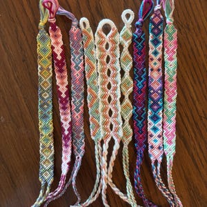 May include: A collection of handmade friendship bracelets in various colors, including pink, blue, yellow, and green. Each bracelet features intricate woven patterns and knotted ends. The bracelets are displayed on a wooden surface.
