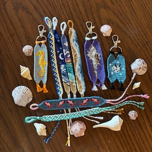 May include: A collection of woven macrame keychains and bracelets featuring various patterns including a yellow seahorse, a night sky, a shell, and geometric designs. The items are crafted with colorful thread and include metal lobster clasps.