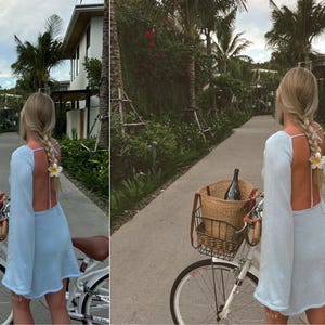 May include: A woman in a light blue dress rides a white bicycle down a paved path lined with palm trees. She has a wicker basket on the front of the bike with a bottle of wine inside.