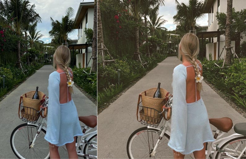 May include: A woman in a light blue dress rides a white bicycle down a paved path lined with palm trees. She has a wicker basket on the front of the bike with a bottle of wine inside.