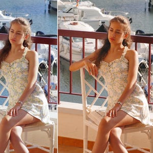 May include: A young person wearing a white floral dress with a ruffled neckline sits on a white wicker chair. The person is looking off to the side and has long brown hair. The chair is on a balcony overlooking a marina with boats in the water.