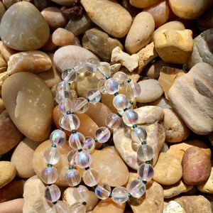 May include: A strand of iridescent clear glass beads with dark green accents, resting on a bed of smooth, light brown stones.