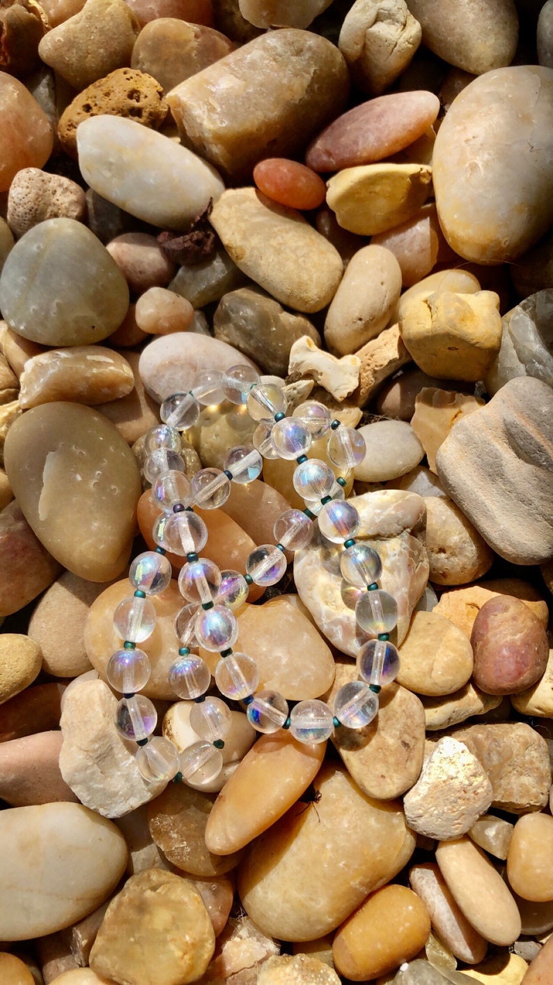 May include: A strand of iridescent clear glass beads with dark green accents, resting on a bed of smooth, light brown stones.