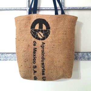 May include: A tan burlap tote bag with navy blue handles. The bag features a black graphic and text that reads "Agroindustrias Un de Mexico S.A. de". The bag is hanging on a metal and wood coat rack.