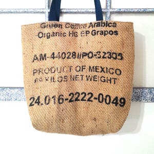 May include: A tan burlap tote bag with dark blue handles. The bag features black text that reads "Green Camo Arabica Organic HG EP Grapos", "AM-44028//PO-32305", "PRODUCT OF MEXICO", "60 KILOS NET WEIGHT", and "24.016-2222-0049".