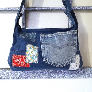 May include: A denim shoulder bag with a patchwork design. The bag features a large denim pocket and several patches of patterned fabric in red, white, and floral prints. The bag's strap is also made of denim.