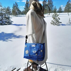 A blue denim crossbody bag with a distressed look and  vintage mother of pearl buttons. The bag has a flap closure.  This is a picture taken outdoors.  The purse is worn by a manikin in a white blouse.