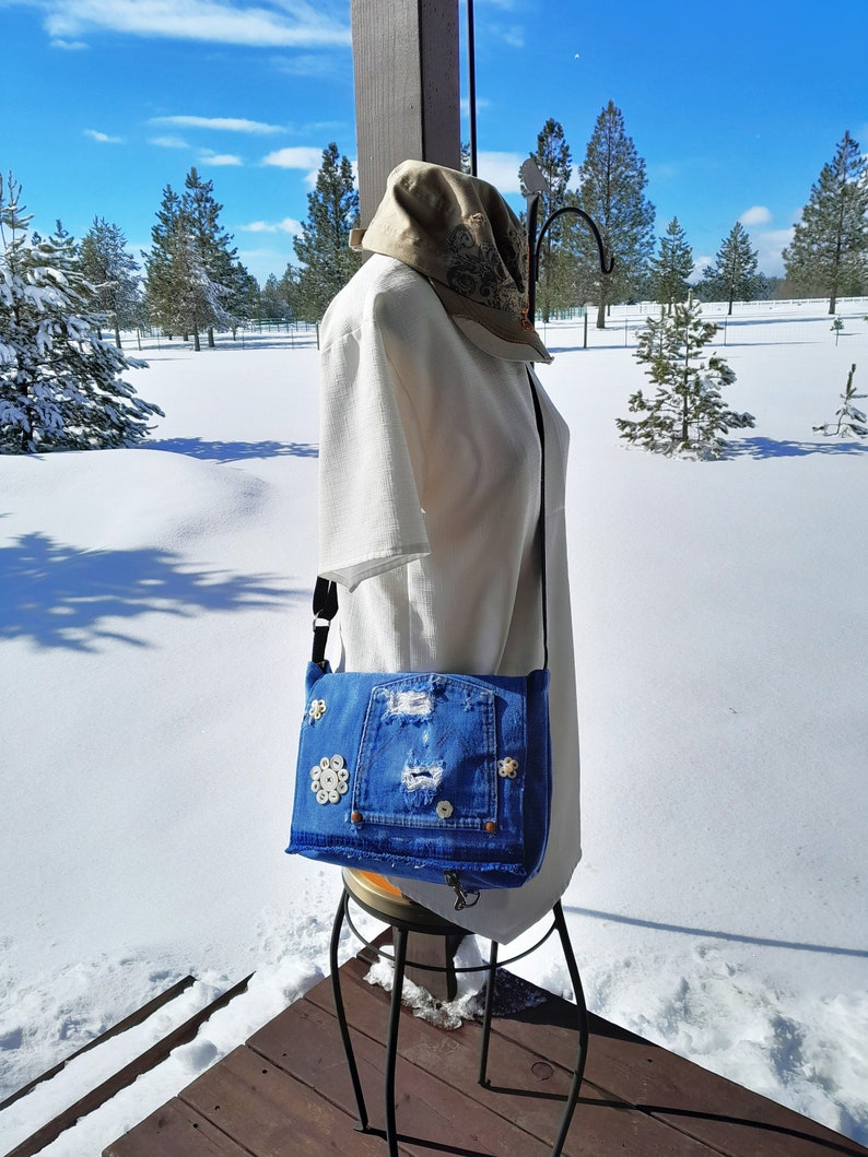A blue denim crossbody bag with a distressed look and  vintage mother of pearl buttons. The bag has a flap closure.  This is a picture taken outdoors.  The purse is worn by a manikin in a white blouse.