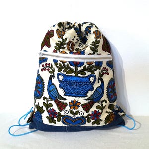 May include: A drawstring backpack with a white background and a colorful floral and bird pattern. The backpack has a blue denim base, a white zipper, and blue drawstring cords. The pattern includes blue birds, flowers, and a blue vase.