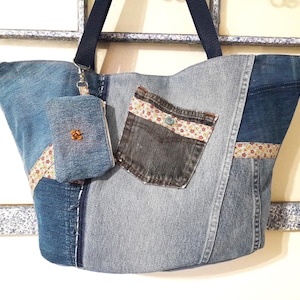 May include: A large denim tote bag with a smaller denim pouch attached. The tote bag is made from different shades of blue denim and has a floral patterned fabric trim. The pouch is also made from denim and has a brown button.