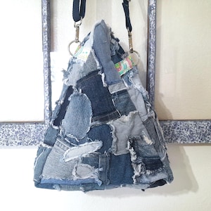 May include: A denim patchwork tote bag with a triangular shape. The bag is constructed from various shades of blue denim, creating a textured, multi-toned appearance. It features a black strap and silver-toned metal hardware for attaching the strap.