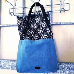 A blue denim cross body bag with a black floral fabric lining.  There is a hook and loop closure and a snap hook closure.
