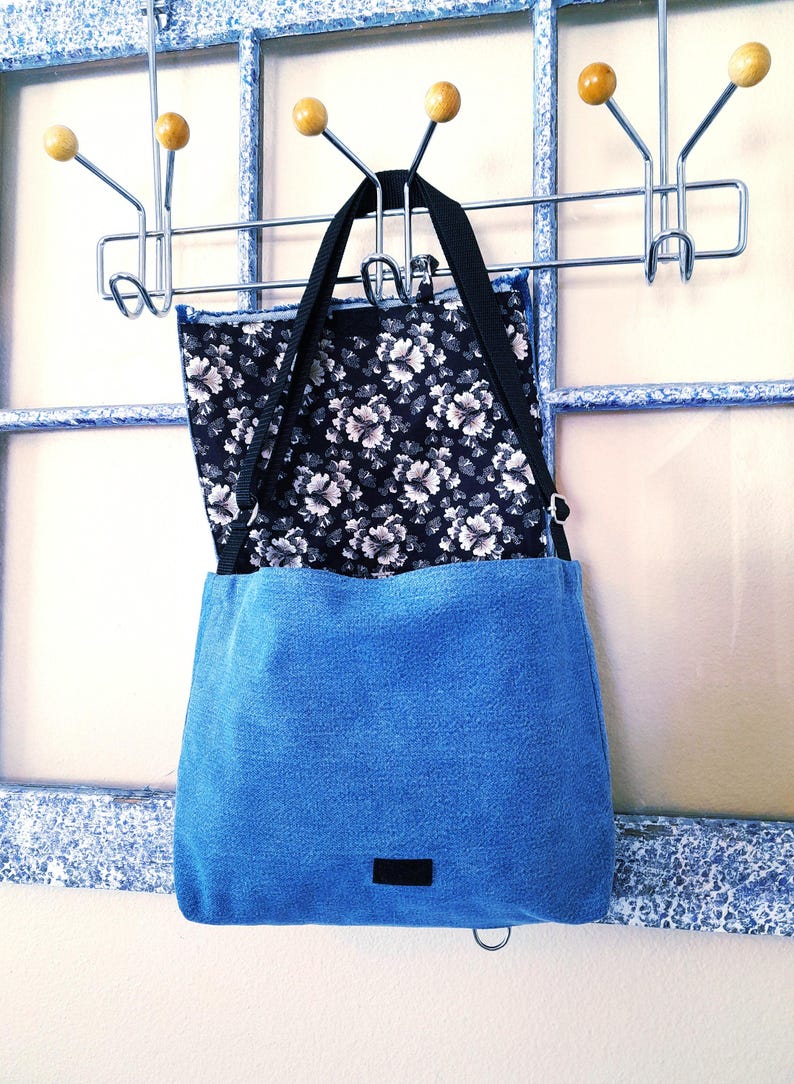A blue denim cross body bag with a black floral fabric lining.  There is a hook and loop closure and a snap hook closure.