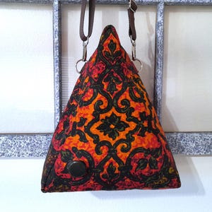 May include: A triangular-shaped bag with a vibrant pattern in shades of red, orange, and black. The bag features a decorative floral design and a black button. It has a brown strap and silver hardware for hanging.