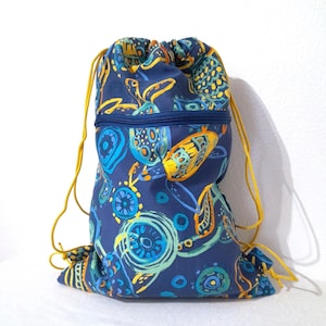 May include: A navy blue drawstring backpack with a colorful pattern of turtles and abstract shapes in shades of blue, yellow, and green. The backpack has a zippered front pocket and yellow drawstrings.