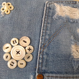 A close-up of a blue denim fabric purse flap with a cluster of white mother of pearl buttons. The buttons are arranged in a circular pattern, with a larger button in the center and smaller buttons surrounding it. The denim fabric has a distressed look, with some areas of the fabric frayed raw edges.
