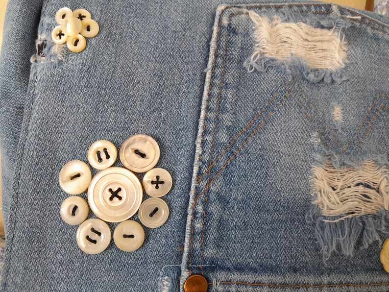 A close-up of a blue denim fabric purse flap with a cluster of white mother of pearl buttons. The buttons are arranged in a circular pattern, with a larger button in the center and smaller buttons surrounding it. The denim fabric has a distressed look, with some areas of the fabric frayed raw edges.