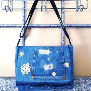 A blue denim crossbody bag with an adjustable strap. The bag has distressed details and  vintage mother of pearlbuttons. The bag has a silver a hook clasp.