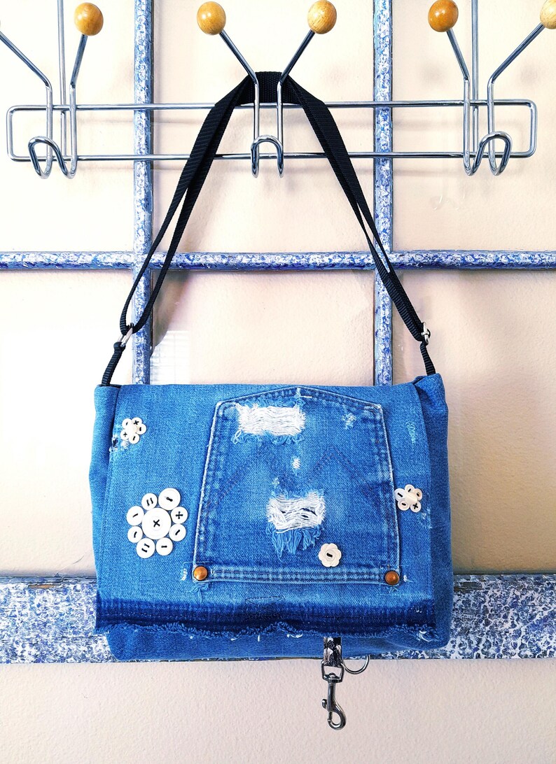A blue denim crossbody bag with an adjustable strap. The bag has distressed details and  vintage mother of pearlbuttons. The bag has a silver a hook clasp.