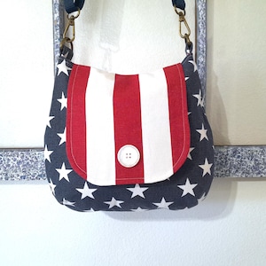 May include: A patriotic-themed shoulder bag with a flap closure. The bag features a navy blue fabric with white stars and a flap with red and white stripes. A white button secures the flap. The bag has a navy blue strap.
