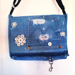 A blue denim crossbody bag with a purse flap. The bag is decorated with vintage mother of pearl buttons. The bag has a distressed look with frayed edges and a large up side down pocket on the front.