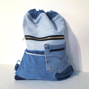 May include: A drawstring backpack made from repurposed denim. The bag features a variety of blue denim washes, a black zipper, and a denim pocket. The bag has blue drawstrings and a denim pocket with a black zipper.