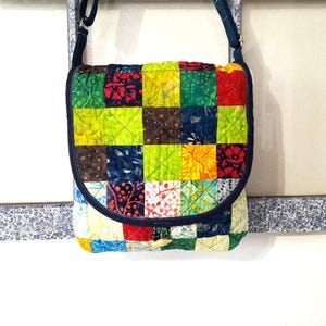 May include: A colorful patchwork crossbody bag with a flap closure. The bag features a vibrant mix of squares in various colors and patterns, including red, green, yellow, and blue. It has a dark blue strap and trim, and a wooden button closure.