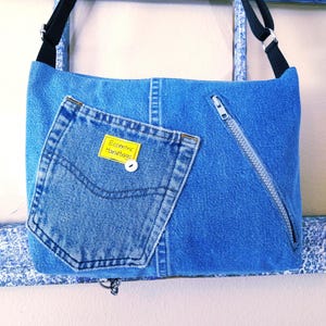 The back of a blue denim crossbody bag with a zippered pocket and a repurposed jeans pocket. The bag has a yellow tag that reads 'Eccentric Handbags'.