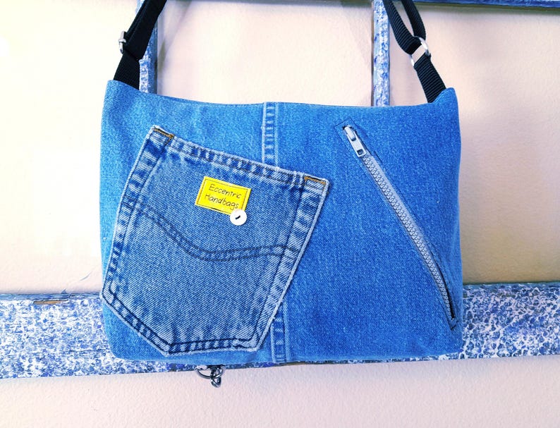 The back of a blue denim crossbody bag with a zippered pocket and a repurposed jeans pocket. The bag has a yellow tag that reads 'Eccentric Handbags'.