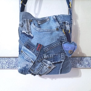 May include: A denim patchwork shoulder bag, crafted from recycled blue jeans. The bag features multiple pockets and a heart-shaped charm. The strap is dark blue, and the bag is set against a white background.