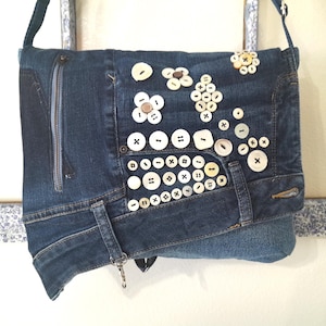 May include: A blue denim messenger bag with a zippered pocket and a flap closure. The bag is decorated with white buttons in various shapes and sizes.
