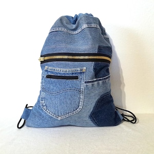 May include: A blue denim drawstring backpack with a black zipper and a pocket. The bag is made from recycled denim and has a black drawstring closure. The zipper has a gold-colored pull.