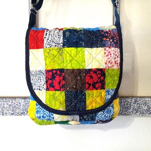 May include: A colorful patchwork crossbody bag with a flap closure. The bag features a variety of fabric squares in red, blue, green, and brown, stitched together in a grid pattern. It has a dark blue strap and trim, and a wooden button closure.