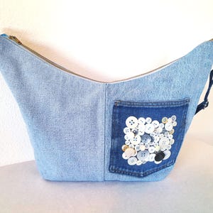 May include: Light blue denim shoulder bag with a zipper closure. The bag features a dark blue denim pocket adorned with an assortment of white, gray, and black buttons. A silver-toned zipper and hardware complete the design.