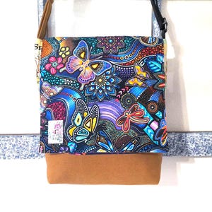 May include: A crossbody bag with a colorful butterfly and floral pattern on a dark blue background. The bag has a brown suede-like bottom and a brown strap. A small white label with a floral design is visible.