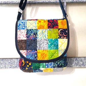 May include: A colorful patchwork crossbody bag with a flap closure. The bag features a variety of patterned squares in shades of blue, yellow, green, and red. It has a dark blue strap and trim, and a silver metal buckle.