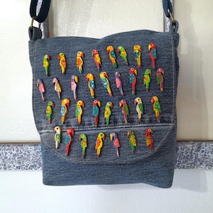 May include: A blue denim crossbody bag featuring a flap decorated with numerous small, colorful wooden parrot-shaped buttons. The parrots are painted in various bright shades including red, yellow, green, and blue, arranged in rows across the front.