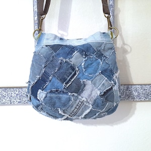 May include: A patchwork denim crossbody bag with a brown strap and brass-colored hardware. The bag is made from various shades of blue denim squares, creating a textured, unique design. The bag hangs against a white wall.