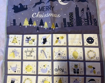 Op de afbeelding: Een grijze kerst adventskalender met 24 genummerde vakken. De bovenkant toont een stadsgezicht, de kerstman in een slee en de woorden "Merry Christmas". Elke zak heeft een feestelijke afbeelding, waaronder klokken, sterren en ornamenten.