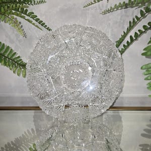 May include: A clear, cut-glass bowl with intricate starburst and geometric patterns. The bowl is round with a scalloped edge, reflecting light and creating a sparkling effect. Green fern fronds frame the bowl, adding a natural contrast.