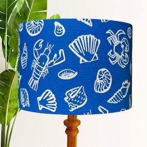 May include: A blue lampshade with a white nautical pattern featuring seashells, a lobster, and a crab. The lampshade is cylindrical and sits on a wooden lamp base. The background includes a green plant.