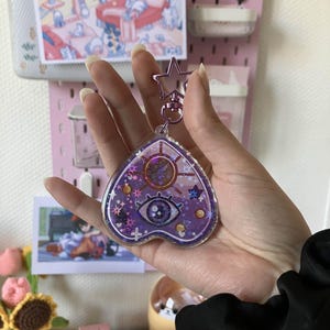 May include: Purple heart-shaped keychain with a glitter-filled resin design. The design features a large eye, stars, and other celestial symbols.