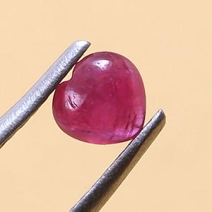 May include: A heart-shaped, polished ruby gemstone, held by silver tweezers. The ruby is a deep, saturated red color, with a smooth, rounded surface. The background is a light tan color.