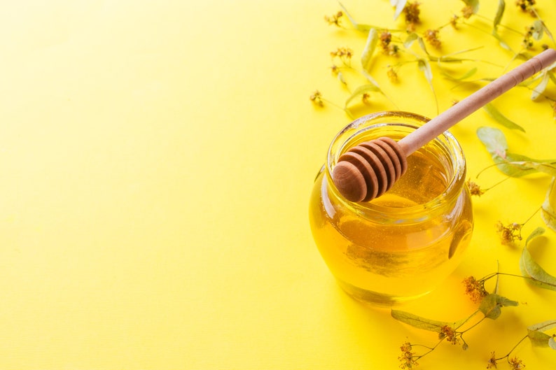May include: A glass jar of honey with a wooden honey dipper on a yellow background. Linden blossoms are scattered around the jar.