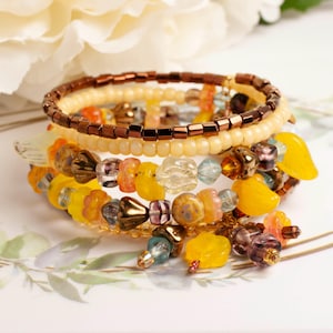 Könnte beinhalten: Ein mehrreihiges Wickelarmband mit einer Vielzahl von Perlen in Gelb-, Orange-, Braun- und Klarglastönen. Das Design umfasst metallisch braune Saatperlen, undurchsichtige gelbe blumenförmige Perlen und durchscheinende bernsteinfarbene Akzente.