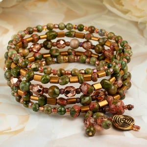 May include: A multi-strand wrap bracelet featuring a combination of round green and pink unakite beads, metallic bronze cylindrical spacers, and faceted copper-toned glass beads. The bracelet ends with a dangling charm featuring a spiral design.