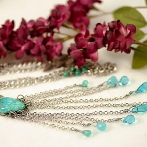 May include: A silver-toned necklace with a large, turquoise-colored stone pendant. The pendant is surrounded by a decorative metal frame. The necklace features multiple strands of chain with teardrop-shaped, light blue beads. Dark red flowers and green leaves are in the background.
